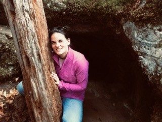 Sarah squats in front of a cave and leans on a tree trunk